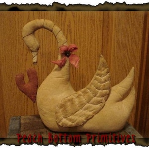 Könnte beinhalten: Ein handgefertigter primitiver Stoffschwan mit einem roten Herzen und einer roten Blume auf dem Kopf. Der Schwan sitzt auf einer Holzoberfläche.