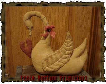 May include: A handmade primitive style fabric swan with a red heart and a red flower on its head. The swan is sitting on a wooden surface.