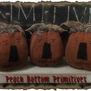 May include: Three brown fabric pumpkins with stitched details and black eyes. The pumpkins are sitting on a white shelf with the text "Peach Bottom Primitives" written in red.