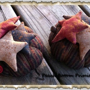 May include: Two handmade fabric pumpkins decorated with stars in shades of brown, red, and white. The pumpkins are sitting on a wooden surface. The text "Peach Bottom Primitives" is visible in the bottom right corner of the image.