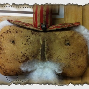 May include: A handcrafted Santa Claus face decoration in a rustic style. The face is light brown with dark brown spots, a white beard, and a red and white striped hat with a small bell.