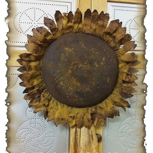 May include: A rustic, brown and yellow sunflower made of fabric with a metal centre. The sunflower is hanging on a wall with a white background. The text "Peach Bottom Primitives" is visible at the bottom of the image.