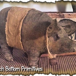 May include: A brown and tan handmade pig figurine with a bell attached to its neck. The pig is standing on a checkered fabric.