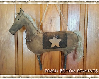 May include: A brown and white primitive horse with a star on its back. The horse is hanging from a string and has a wooden base. The text "Peach Bottom Primitives" is visible at the bottom of the image.