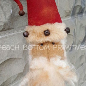 May include: A handmade felt Christmas gnome with a red hat, white beard, and brown felt accents. The gnome is wearing a brown felt hat with a bell on top. The gnome is hanging on a brick wall.