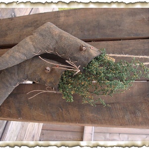 Puede incluir: Dos botas de tela marrón con campanas colgando de una tabla de madera rústica. Las botas están llenas de vegetación seca y atadas con cuerda. El texto "Peach Bottom Primitives" es visible en la parte inferior de la imagen.
