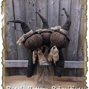 May include: Three handmade primitive witch dolls with black hats and burlap accents. The dolls are arranged in a glass jar with sticks for legs. The dolls are standing in front of a wooden fence.