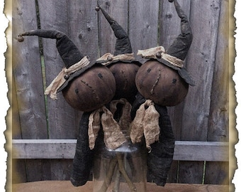 May include: Three handmade primitive witch dolls with black hats and burlap accents. The dolls are arranged in a glass jar with sticks for legs. The dolls are standing in front of a wooden fence.