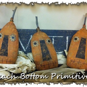 May include: Three handmade primitive fabric pumpkins with stitched faces and black eyes. The pumpkins are arranged in a row on a wooden surface with the text "Peach Bottom Primitives" and the year "2014" visible.