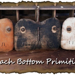 May include: Three handmade primitive pumpkin decorations in orange, black, and white. Each pumpkin has a simple face with eyes, a nose, and a mouth. The pumpkins are arranged in a row on a wooden surface. The text "Peach Bottom Primitives" is visible below the pumpkins.