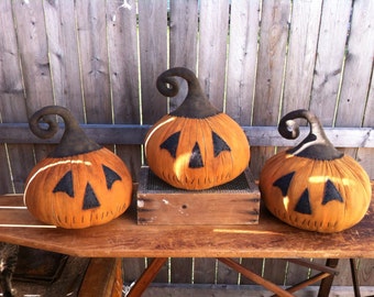 May include: Three handmade felt pumpkins with black stems and stitched facial features. The pumpkins are sitting on a wooden table in front of a wooden fence.