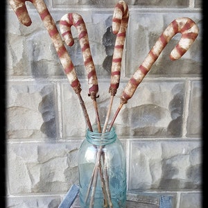 Puede incluir: Cuatro decoraciones de bastón de caramelo rayadas de rojo y blanco en un frasco de vidrio transparente con un tinte azul. Los bastones de caramelo están dispuestos en un jarrón con palos marrones. El frasco está sentado sobre una superficie de madera.