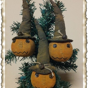 Puede incluir: Tres adornos de calabaza de fieltro hechos a mano con sombreros de bruja. Las calabazas son de color naranja con detalles pintados y los sombreros son marrones con una campana en la parte superior. Los adornos están colgados de un árbol de Navidad artificial verde.