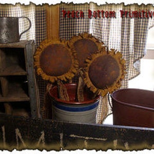 May include: Three rustic brown and orange fabric sunflowers with frayed edges in a white and blue metal bucket. The sunflowers are on a wooden shelf with a sign that says "Prick Bottom Primitives".