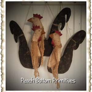May include: Three black and white primitive rooster ornaments with red combs and wattles hanging from a white wall. The ornaments are made of fabric and have a rustic, vintage look. The text "Peach Bottom Primitives" is visible at the bottom of the image.