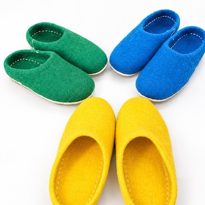 May include: Three pairs of felted wool slippers arranged in a circle. One pair is vibrant green, one pair is bright blue, and one pair is golden yellow. Each slipper features a rounded toe, an open top, and visible white stitching along the inner rim.