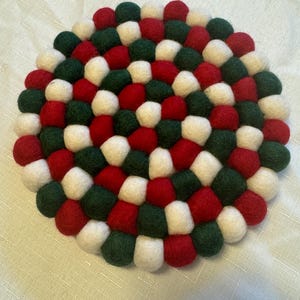May include: A round trivet made of felt balls in red, white, and green. The trivet is composed of numerous small, tightly packed felt balls, creating a textured surface. The color scheme suggests a festive or holiday theme.