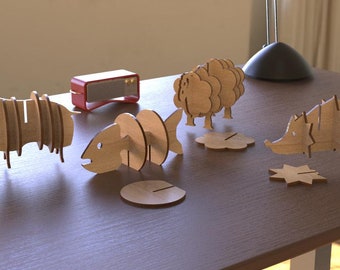 Puede incluir: Un conjunto de cinco figuritas de animales de madera, que incluyen un hipopótamo, un pez, una oveja y un erizo. Cada figurita está hecha de múltiples capas de madera y tiene un diseño único. Las figuritas se muestran en una mesa de madera.