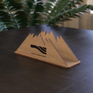 Puede incluir: Servilletero de madera con diseño de picos montañosos. El soporte, de madera marrón claro, presenta un gráfico negro y el texto "WOODEN NAPKIN HOLDER". Se encuentra sobre una superficie marrón oscuro.