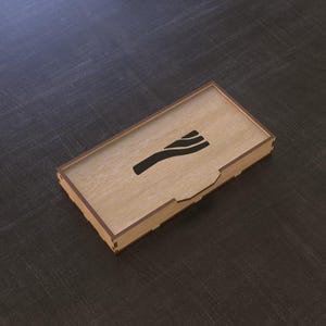 May include: A rectangular wooden box with a laser-cut design on the lid. The box is light brown with a dark brown border and a black graphic. The box is on a dark grey surface.