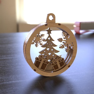 Könnte beinhalten: Weihnachtsbaumschmuck aus Holz mit kreisförmigem Rahmen. Im Inneren sind ein detaillierter Baum, Schneeflocken und verpackte Geschenke ausgeschnitten. Der Schmuck hat eine Schlaufe zum Aufhängen. Das Holz hat eine natürliche, hellbraune Farbe.