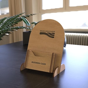 May include: A wooden desk plaque with a business card holder. The plaque has the words "BRAND DESK PLAQUE" and "BUSINESS CARD" printed on it. The plaque is light brown and has a curved top.
