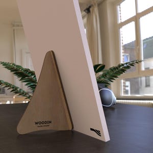 Könnte beinhalten: Ein hölzerner Staffeleiständer mit dreieckiger Stütze und weißer Oberfläche. Der Ständer besteht aus hellbraunem Holz und trägt den Text "WOODIN EASEL STAND". Ein schwarzes Logo ist auf der weißen Oberfläche sichtbar.