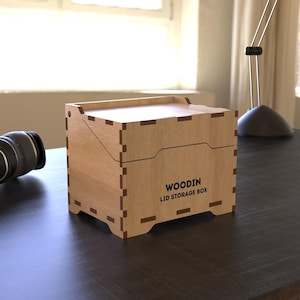 Puede incluir: Una caja de almacenamiento de madera de color claro con el texto "WOODIN LID STORAGE BOX" en la parte delantera. La caja tiene una tapa y está sobre una superficie oscura. Unos auriculares negros están a la izquierda.