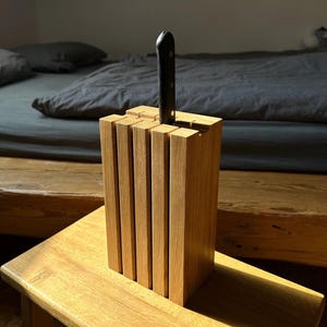 Op de afbeelding: Een houten messenblok met een enkel zwart mes erin. Het blok is gemaakt van lichtgekleurd hout en heeft een rechthoekige vorm met verticale sleuven. Het messenblok staat op een houten oppervlak.