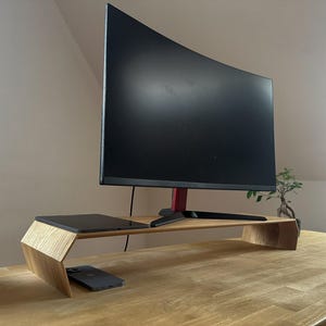 Puede incluir: Un soporte de monitor de madera con una pantalla negra curva y detalles en rojo. Una computadora portátil y un teléfono inteligente están en el soporte. El soporte está sobre un escritorio de madera. Un bonsái y una planta están en el fondo.