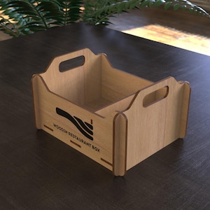 May include: A light brown wooden restaurant box with a rectangular shape and two handles. The box has a black logo and the text "WOODIN RESTAURANT BOX" printed on the side. The box is sitting on a dark brown surface.