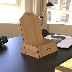 Könnte beinhalten: Ein Schreibtisch-Organizer aus hellem Holz mit schräger Rückseite und rechteckiger Basis. Der Organizer trägt oben den Text "WOODIN" und vorne "AA PROSPECT". Eine schwarze Schreibtischlampe, Kopfhörer und ein offenes Buch befinden sich ebenfalls auf dem Schreibtisch.