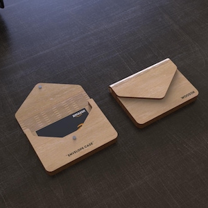 Puede incluir: Dos estuches de madera con forma de sobre, uno abierto y otro cerrado, sobre una superficie oscura. El estuche abierto revela una tarjeta de regalo de Amazon. El texto "ENVELOPE CASE" está impreso en el estuche abierto. El estuche cerrado tiene la palabra "WOODIN".