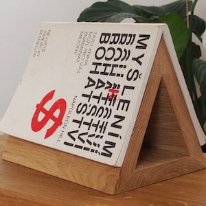 Könnte beinhalten: Ein Bücherständer aus Holz mit einem dreieckigen Design. Der Ständer hält ein Buch mit dem Titel "Denke nach und werde reich" von Napoleon Hill. Der Buchumschlag ist weiß mit schwarzem Text und einem roten Dollarzeichen.