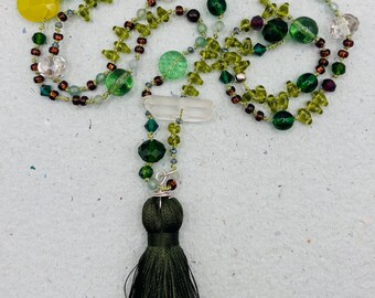 Op de afbeelding: Een lange groene en gele kralenketting met een groene kwast. De ketting heeft verschillende groene, gele en heldere kralen, evenals een hanger van helder kwartskristal.