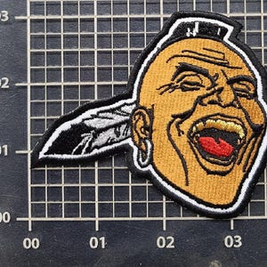 May include: Embroidered patch featuring a Native American chief with a wide open mouth and a feather in his hair. The patch has a black border and is primarily tan, red, and black.