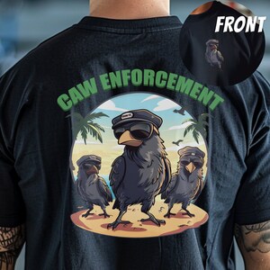 以下が含まれることがあります： 黒いTシャツに、サングラスと帽子をかぶったカラス3羽のグラフィックがプリントされています。カラスの上に緑色の文字で「CAW ENFORCEMENT」と書いてあります。