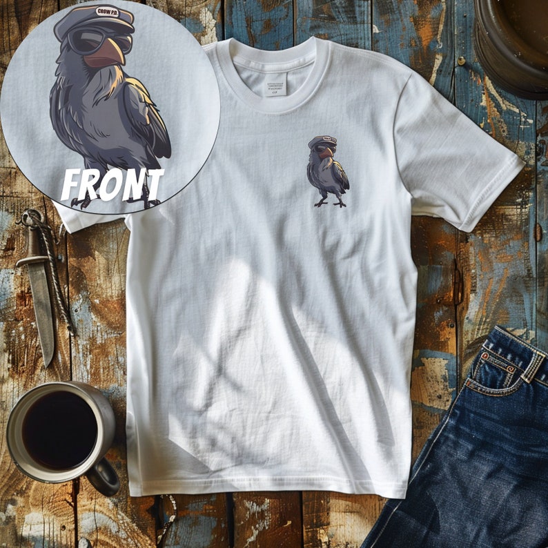 以下が含まれることがあります： 白いTシャツに、船長の帽子とサングラスをかけた漫画の鳥が描かれています。鳥は灰色で、黒いクチバシをしています。Tシャツは木製の表面に平らに置かれています。