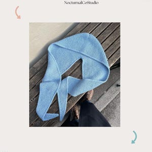 Op de afbeelding: Lichtblauwe gehaakte sjaal met capuchon, gedrapeerd over een houten bank. Het ontwerp van de sjaal omvat een capuchon en lange, vloeiende uiteinden. De tekst "Hooded Scarf" en "NocturnalCoStudio" staat bovenaan de afbeelding.