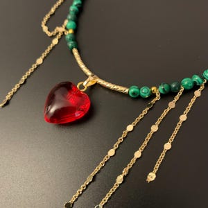 Op de afbeelding: Een gouden ketting met een rode hartvormige hanger en groene kralen. De ketting heeft meerdere gouden kettingen met kleine ronde schakels die eraan hangen.