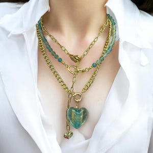 May include: A multi-strand necklace set with gold-coloured chains and teal beaded strands. It features a clear heart-shaped pendant with gold detailing, and a small heart charm. The necklace is worn over a white collared shirt.