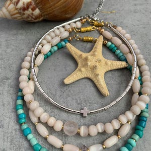 May include: A set of four layered necklaces featuring turquoise and white beads. The necklaces are made with a variety of materials, including metal, shell, and stone. The necklaces are accented with a small cross charm and a pearl.