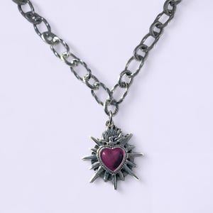 May include: A silver-toned necklace with a large, textured chain. The pendant features a heart-shaped, deep-red center framed by a starburst design. The necklace is displayed against a white background.