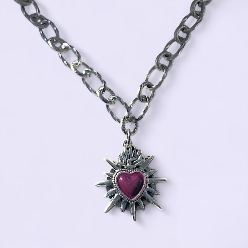 May include: A silver-toned necklace with a large, textured chain. The pendant features a heart-shaped, deep-red center framed by a starburst design. The necklace is displayed against a white background.