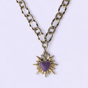 May include: A gold-toned chain necklace with a pendant. The pendant features a purple heart shape surrounded by a gold sunburst design. The chain has large, textured links. The necklace is displayed against a white background.
