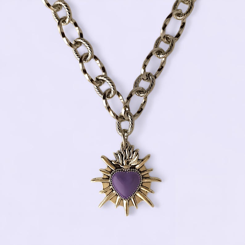 May include: A gold-toned chain necklace with a pendant. The pendant features a purple heart shape surrounded by a gold sunburst design. The chain has large, textured links. The necklace is displayed against a white background.