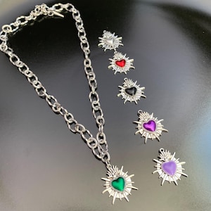 A silver-toned chain necklace with a heart-shaped pendant featuring a green center. Several heart-shaped charms with red, black, purple, and light purple centers are also displayed. The charms have a sunburst design around the heart.