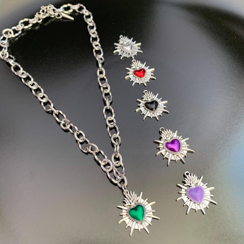 A silver-toned chain necklace with a heart-shaped pendant featuring a green center. Several heart-shaped charms with red, black, purple, and light purple centers are also displayed. The charms have a sunburst design around the heart.