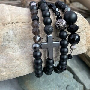 Puede incluir: Cuatro pulseras de cuentas negras apiladas sobre una superficie de madera. Las pulseras presentan cuentas de ónix negro, roca volcánica y cristal. Una pulsera tiene un dije de cruz plateada.