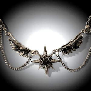 May include: Silver-coloured necklace with a central starburst pendant and a dark gemstone. The necklace has wing-like accents and chain details. The design is gothic-inspired, with a dark, elegant aesthetic.